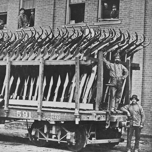 1882 年的照片，三个人站在载满钢犁的 Deere & Co 车厢旁，后面的建筑物中，工厂员工从窗户向外张望