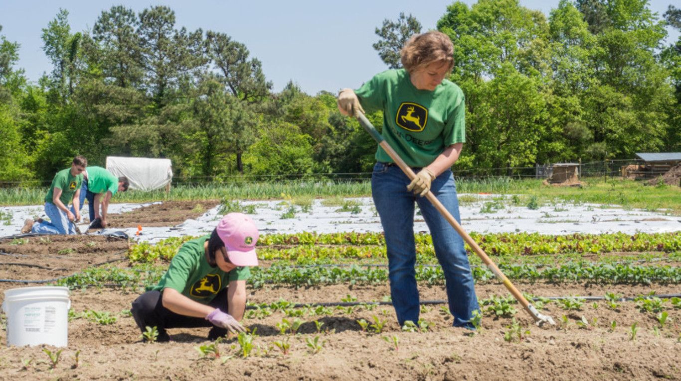 Two women planting and weeding in a large garden with trees and blue sky in the background.